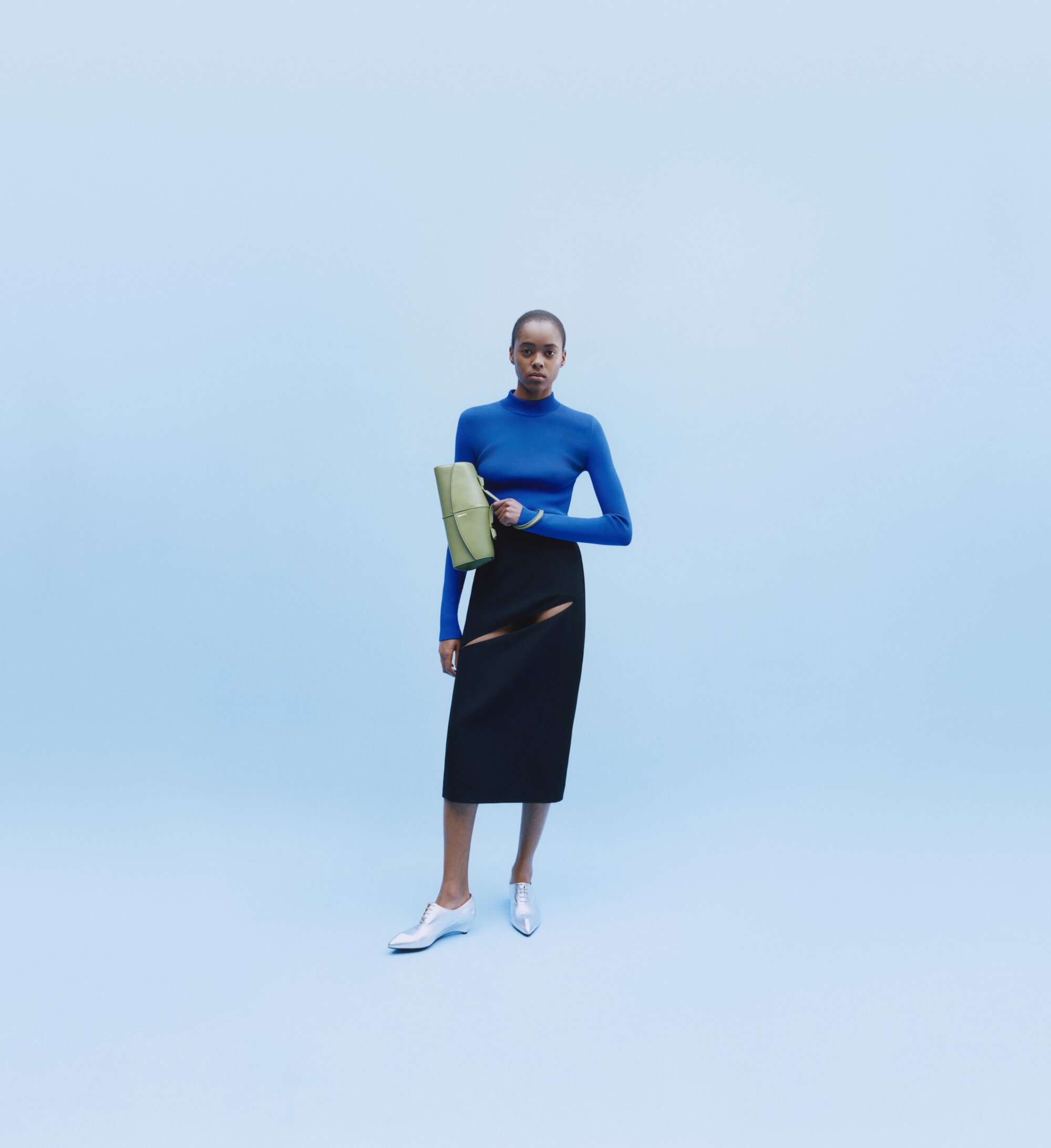 Fashion shot of a Black woman in a blue long-sleeve top and black skirt, holding a green bag, against a light blue backdrop.