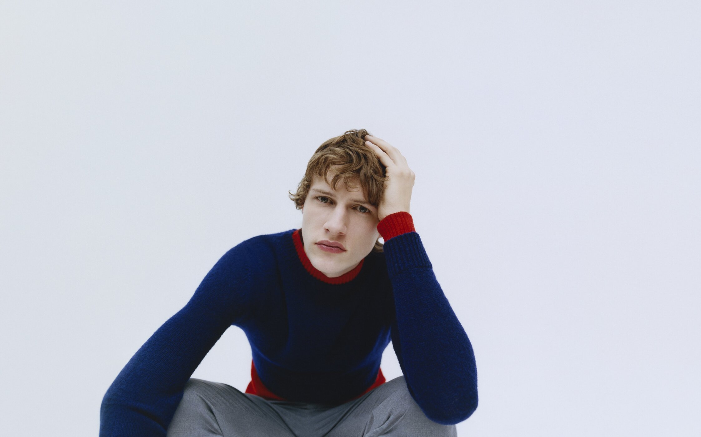 Studio shot of a young man in a blue sweater and gray pants, seated with his hand in his hair against a white background.