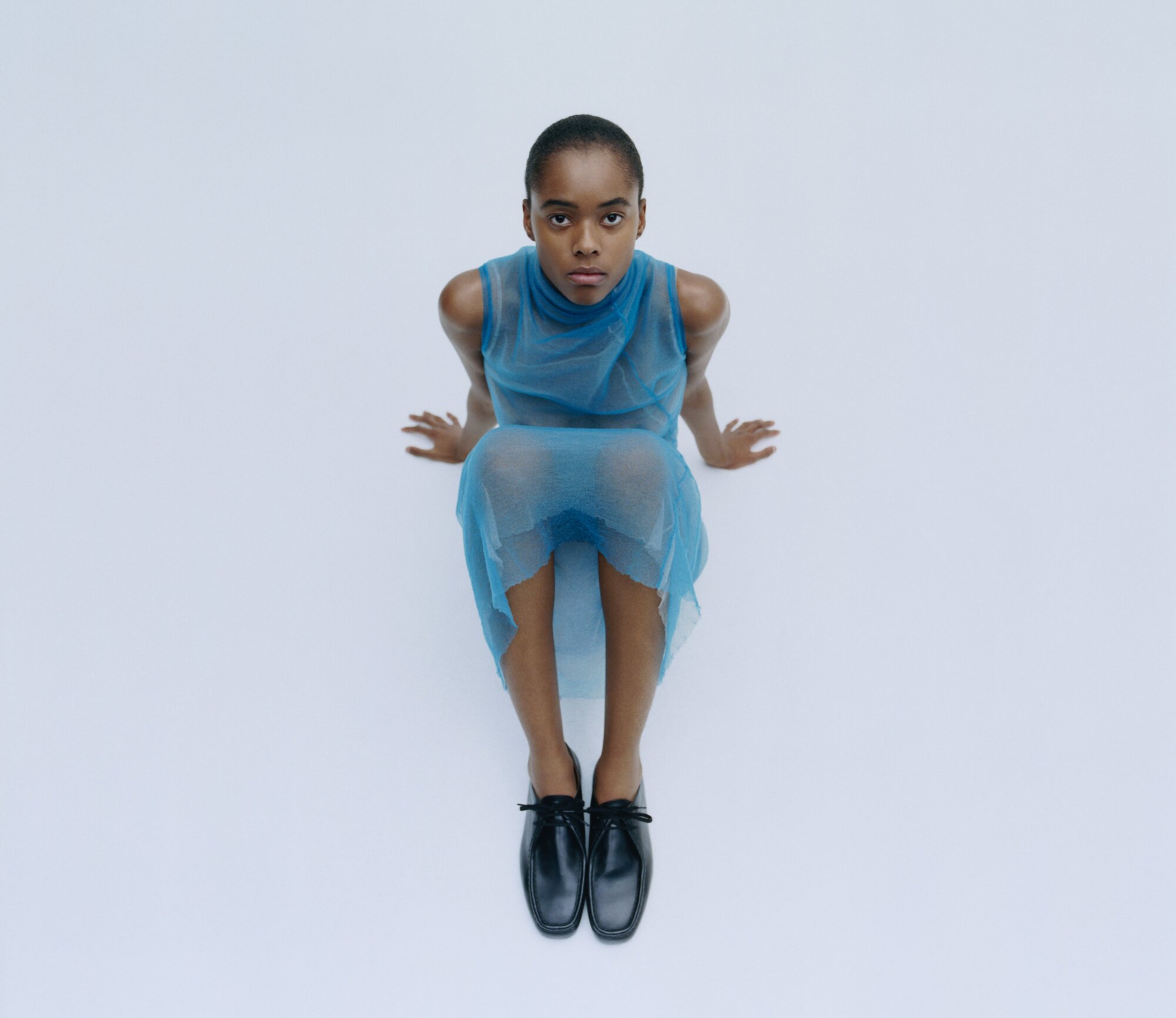 An adult female sits in a photo studio with a high angle view, wearing a blue, sheer dress and black shoes, against a plain white backdrop.
