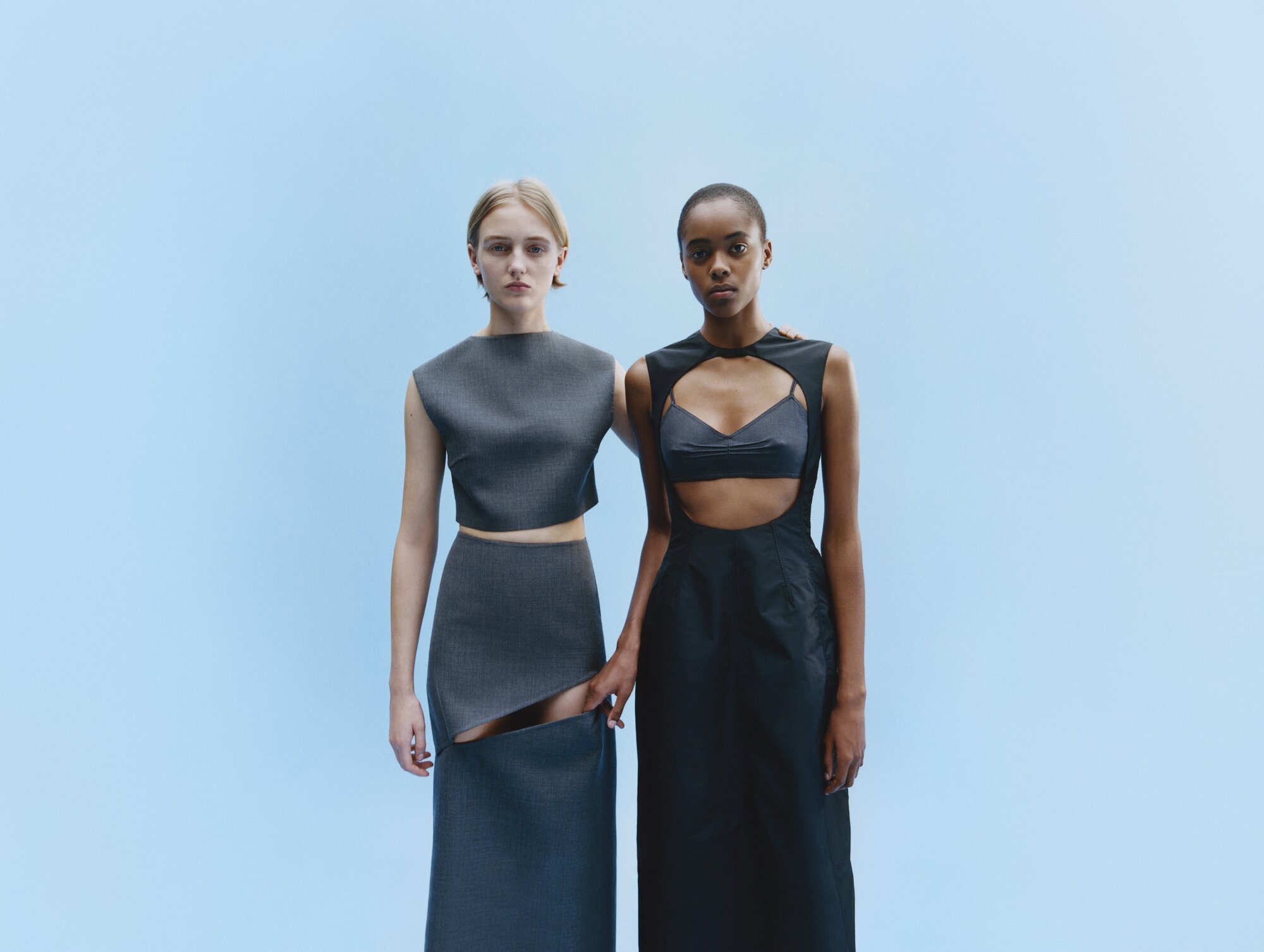 Two women pose in gray and black minimalist dresses against a pale blue backdrop in a studio fashion shot, clean studio lighting.