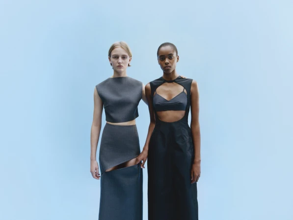Two women pose in gray and black minimalist dresses against a pale blue backdrop in a studio fashion shot, clean studio lighting.
