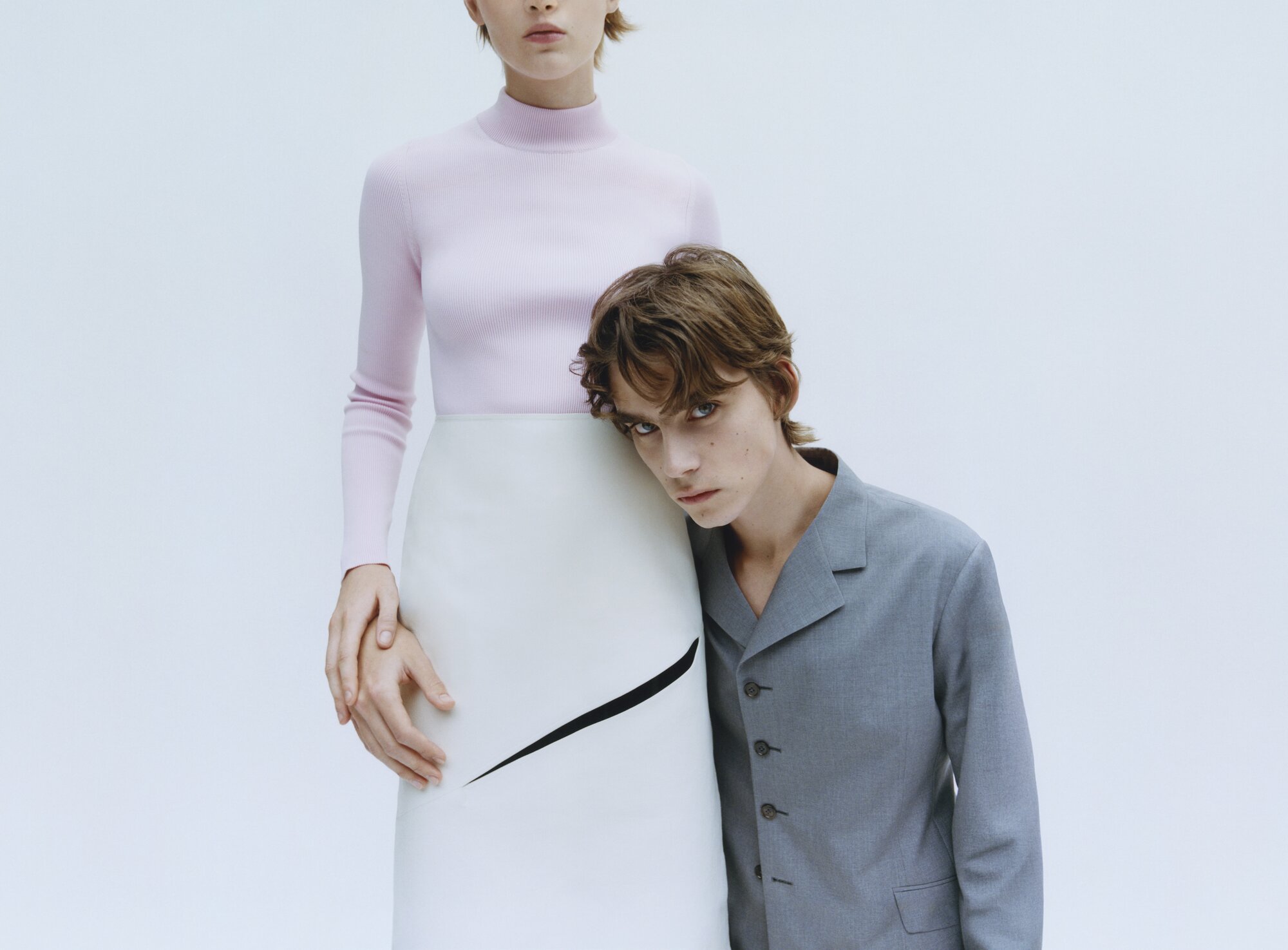 Fashion shot features two models: a woman in a pink top and cream skirt, and a man in a grey suit leaning on the woman's skirt.