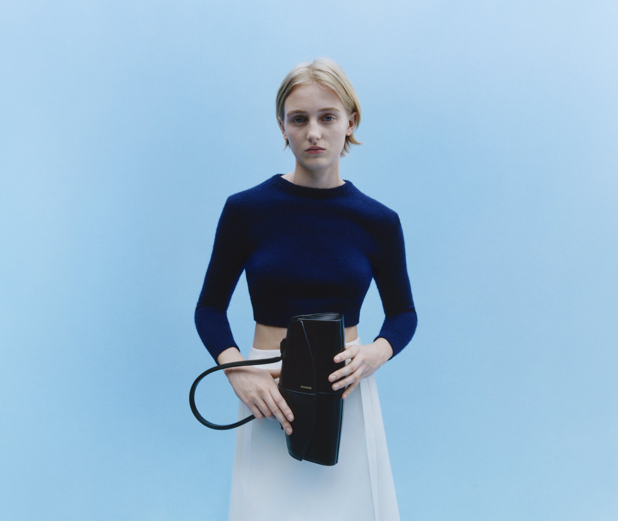 Blonde woman in navy top and white skirt holding a black handbag against a light blue seamless studio background.