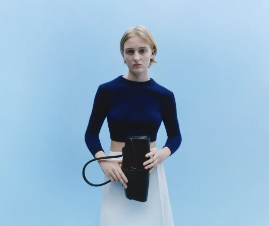Blonde woman in navy top and white skirt holding a black handbag against a light blue seamless studio background.
