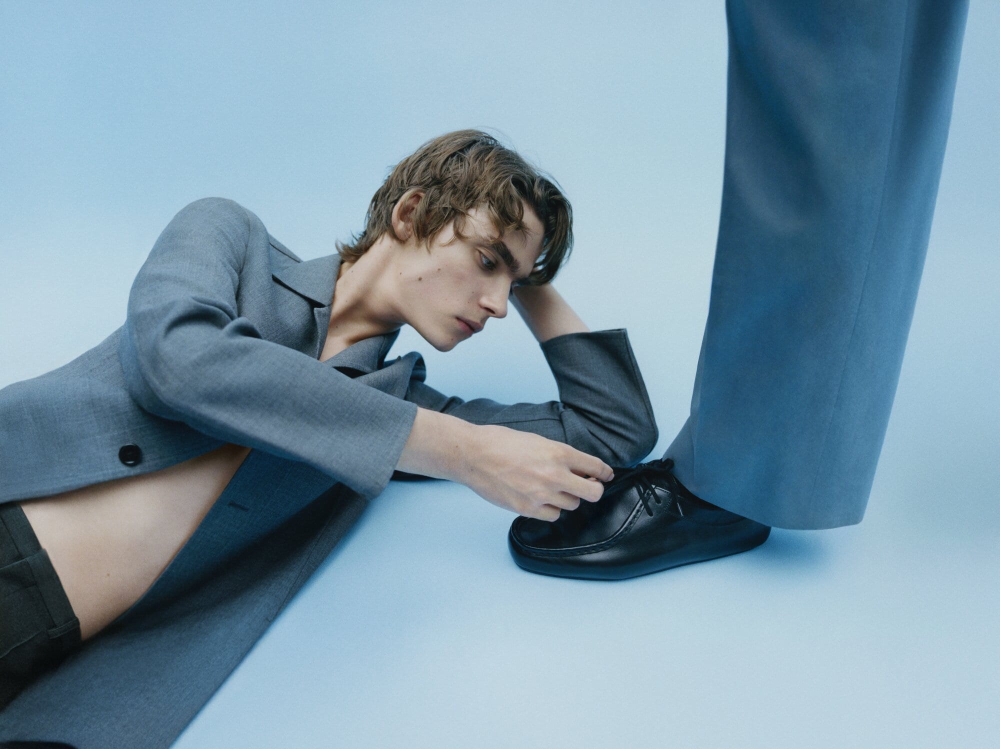 Young adult model in a grey suit jacket poses with a black loafer in a studio shot against a solid light blue backdrop.