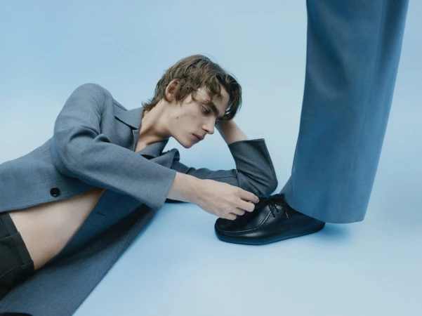 Young adult model in a grey suit jacket poses with a black loafer in a studio shot against a solid light blue backdrop.