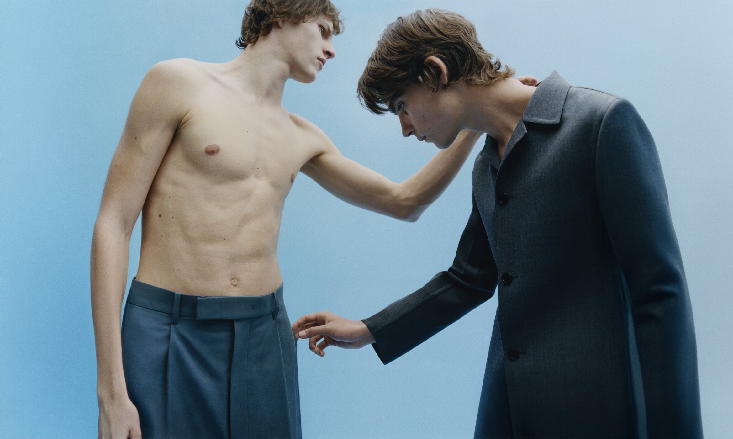 Two young men in a studio setting, one shirtless wearing pants, the other in a dark jacket and pants, against a blue background.