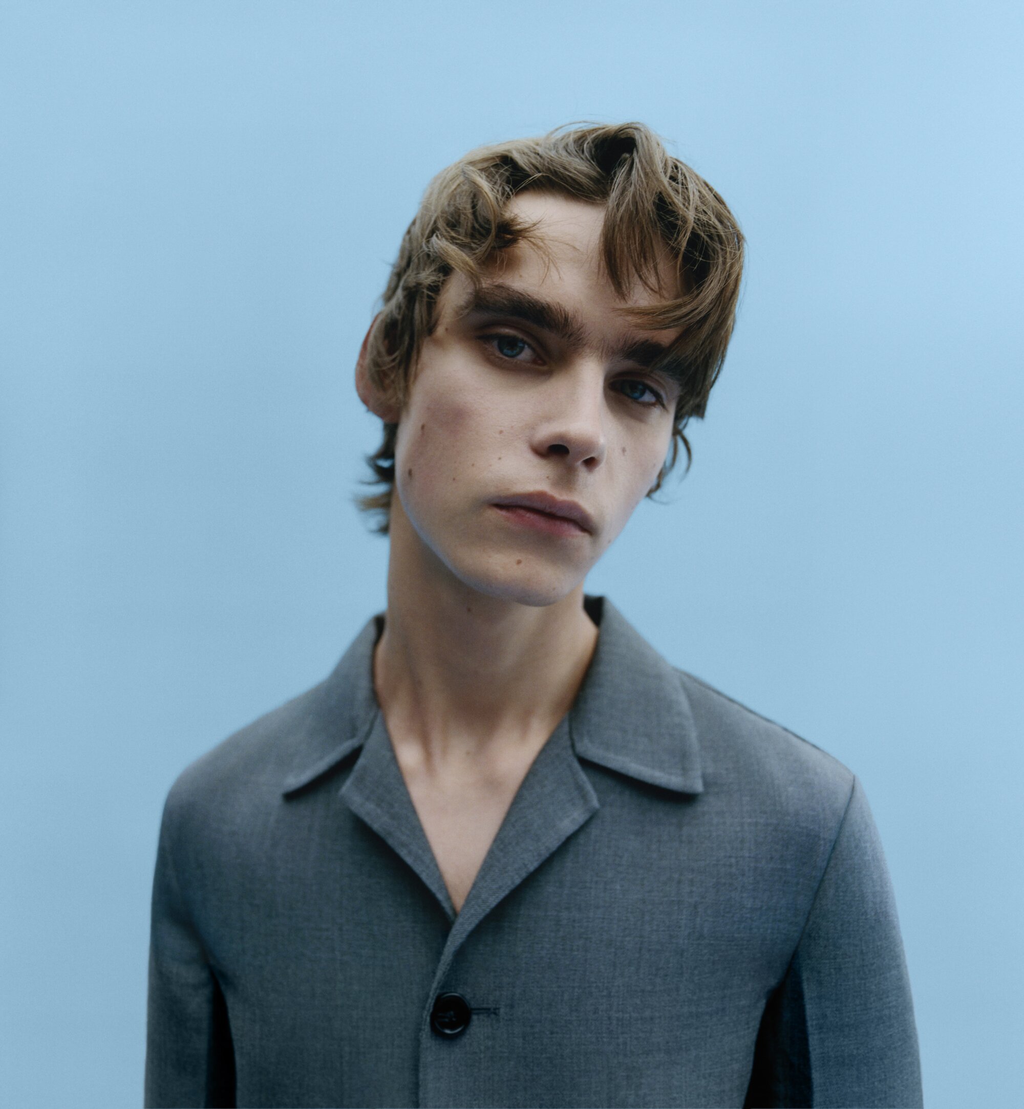 Portrait of a young man wearing a grey button-up collared jacket against a plain light blue background.