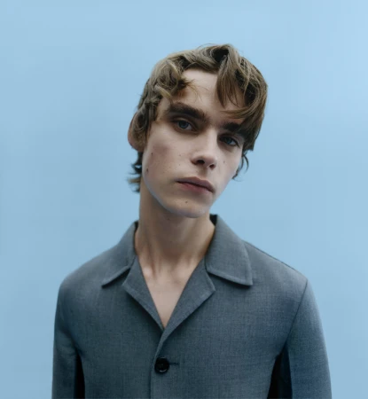 Portrait of a young man wearing a grey button-up collared jacket against a plain light blue background.