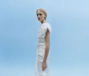 Model in a white textured dress standing sideways against a light blue studio backdrop for the Jil Sander Spring 2026 campaign.