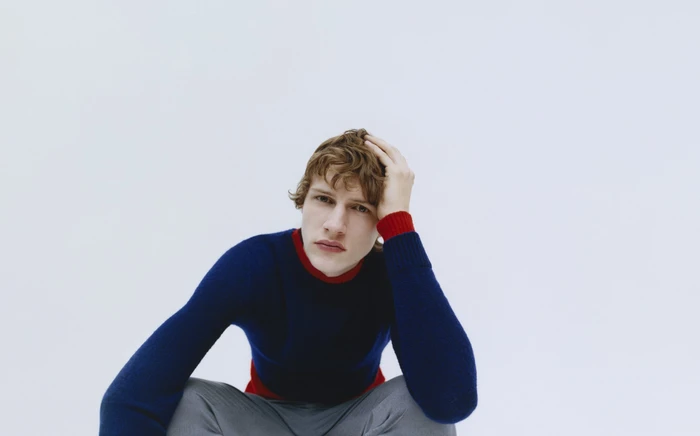 Young man with brown hair wearing a blue sweater sitting against a plain white background