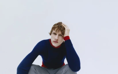Young man with brown hair wearing a blue sweater sitting against a plain white background