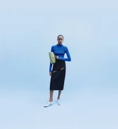 Model in blue top and black skirt holding a green handbag against a blue studio seamless backdrop