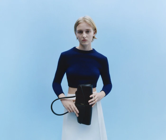 Blonde woman in navy top and white skirt holding a black handbag against a light blue seamless studio background.