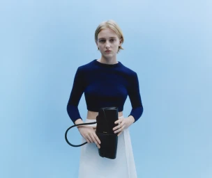 Blonde woman in navy top and white skirt holding a black handbag against a light blue seamless studio background.