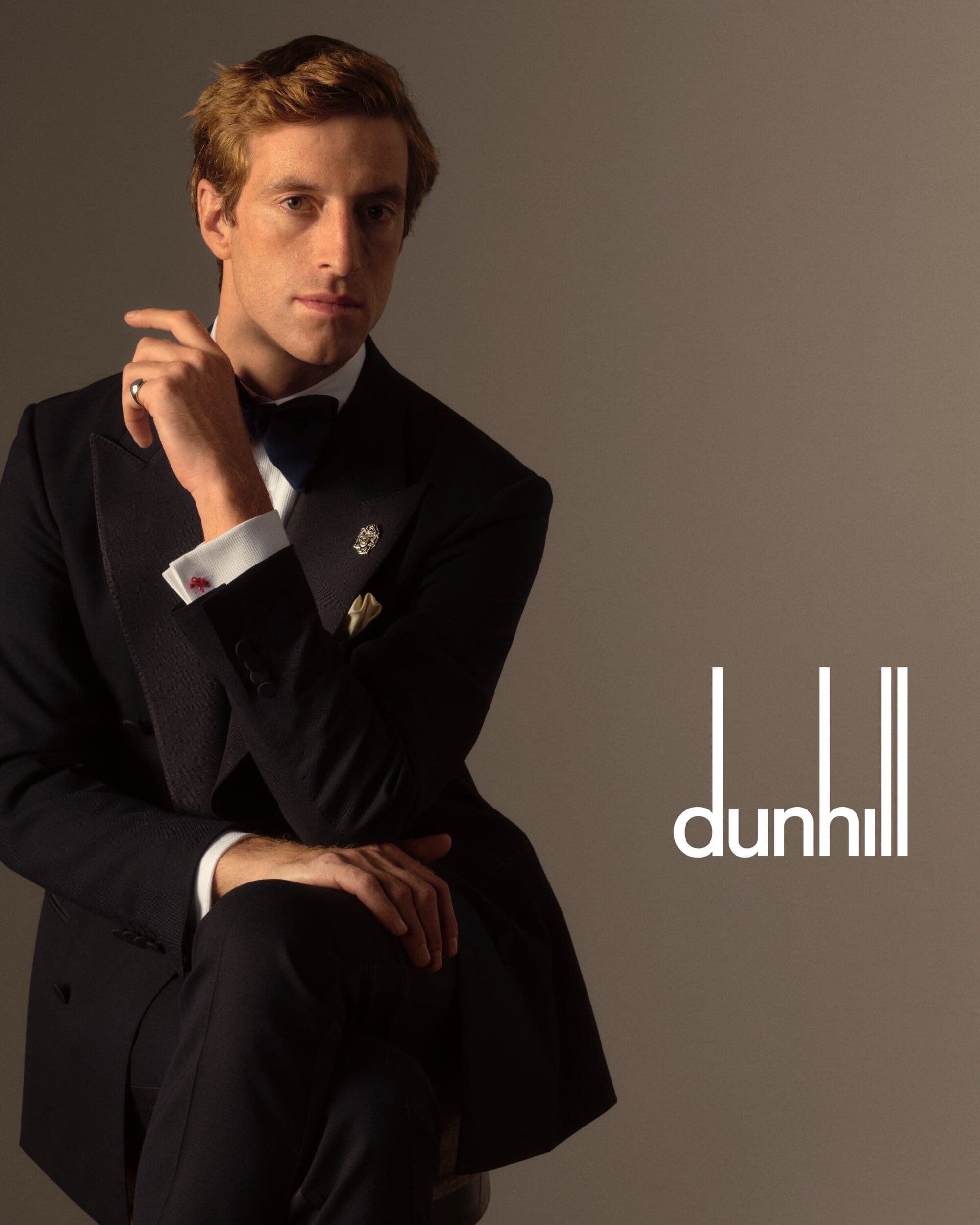 An adult man in a black tuxedo and bowtie poses in a photo studio with the dunhill logo. The man has fair skin and reddish brown hair.