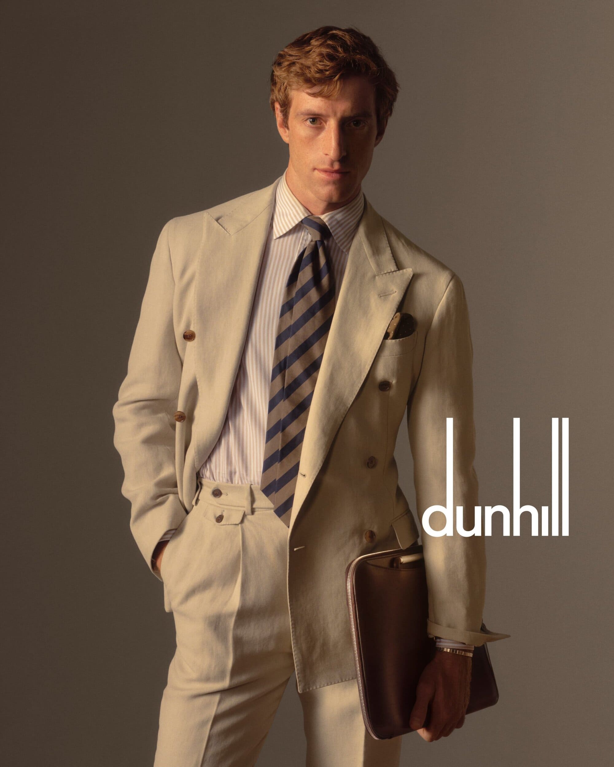 A man in a beige double-breasted suit, striped shirt, and striped tie holds a leather folio in a fashion studio shot.