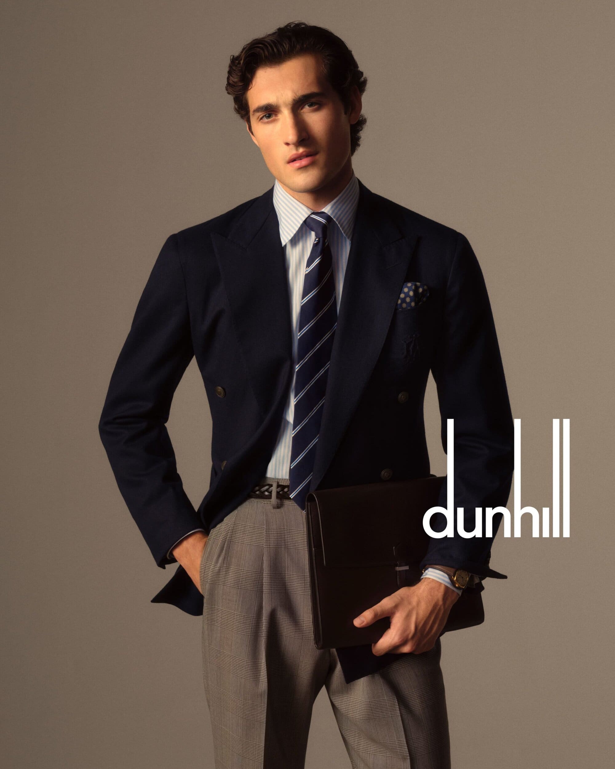 Man in blue blazer, striped shirt and gray pants holding a black briefcase, looking at the camera in a studio setting. Fashion.