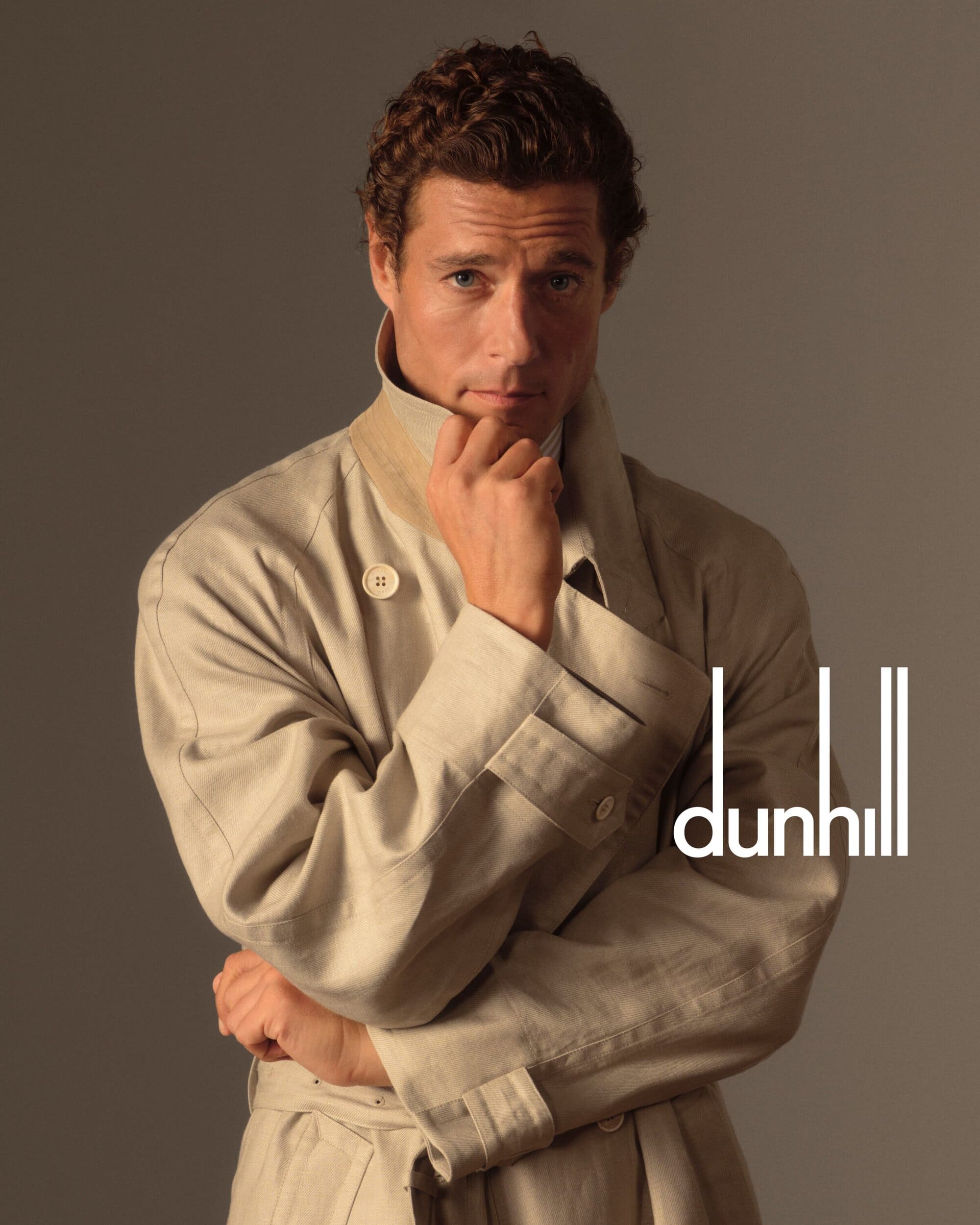 Studio portrait of a handsome male model in a tan trench coat looking at the camera with hand on chin, with Dunhill logo.