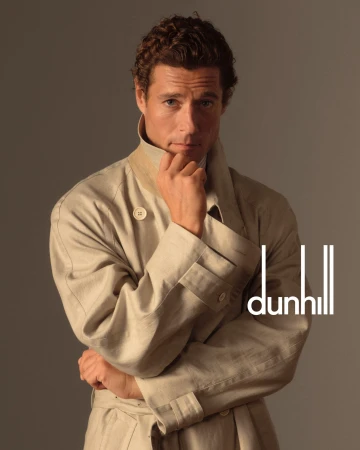 Studio portrait of a handsome male model in a tan trench coat looking at the camera with hand on chin, with Dunhill logo.