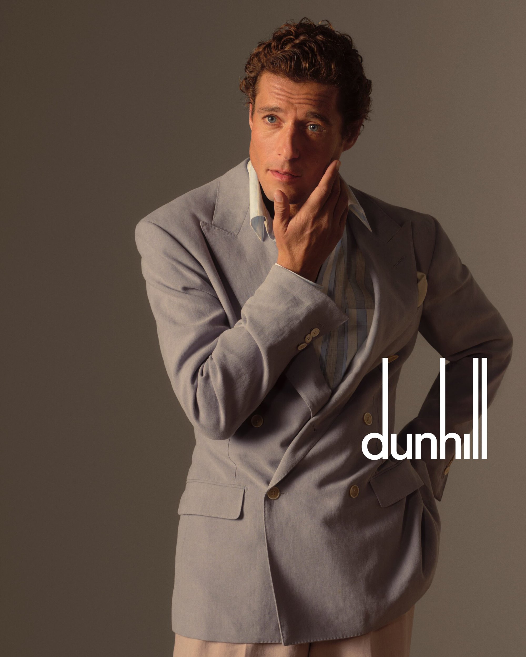 Elegant man in a light grey suit and off-white shirt poses in a studio with a hand on his face, against a neutral grey background.