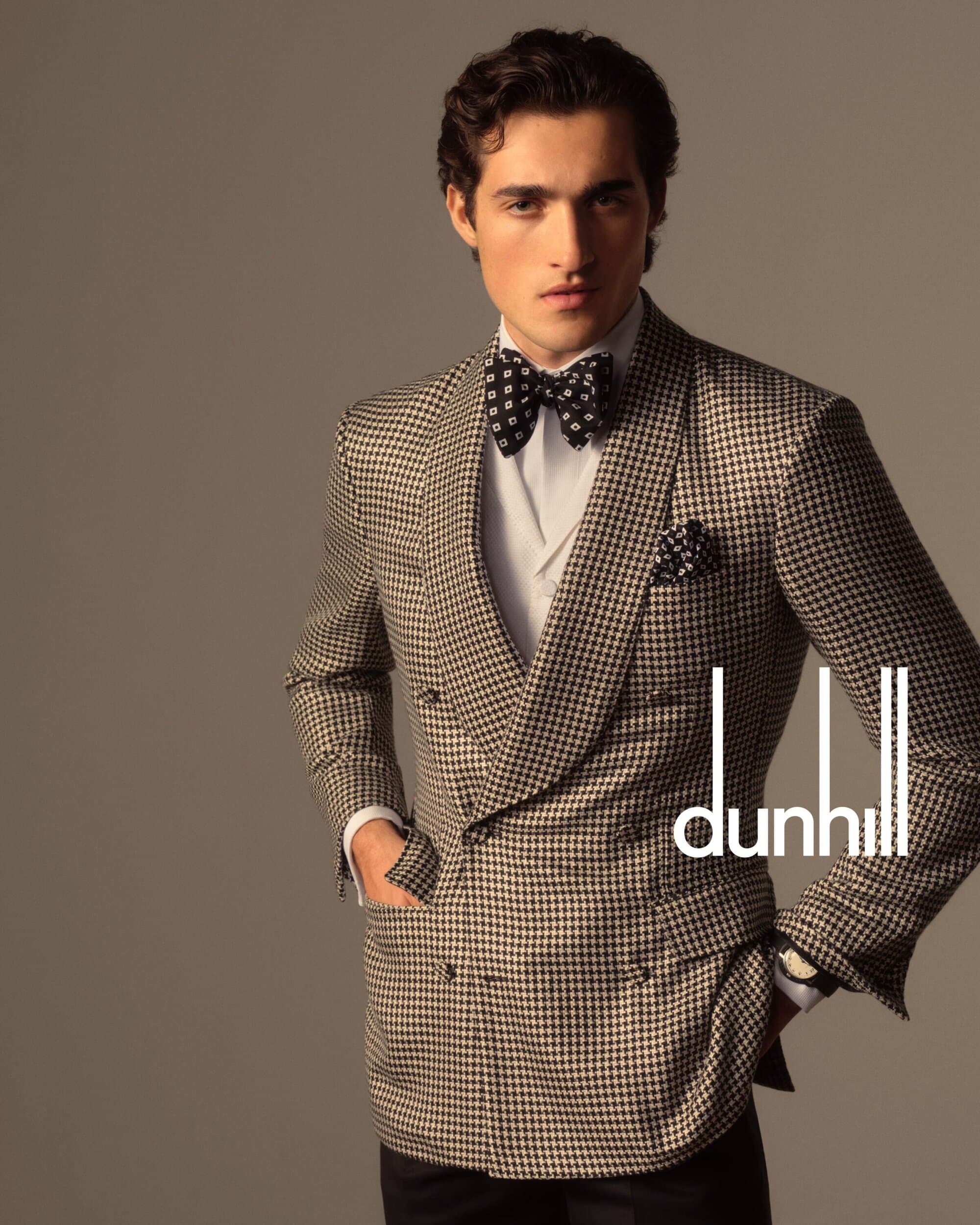 A well-dressed man in a houndstooth suit, vest, and bow tie poses indoors with hands in pockets. Dunhill logo is prominently displayed.