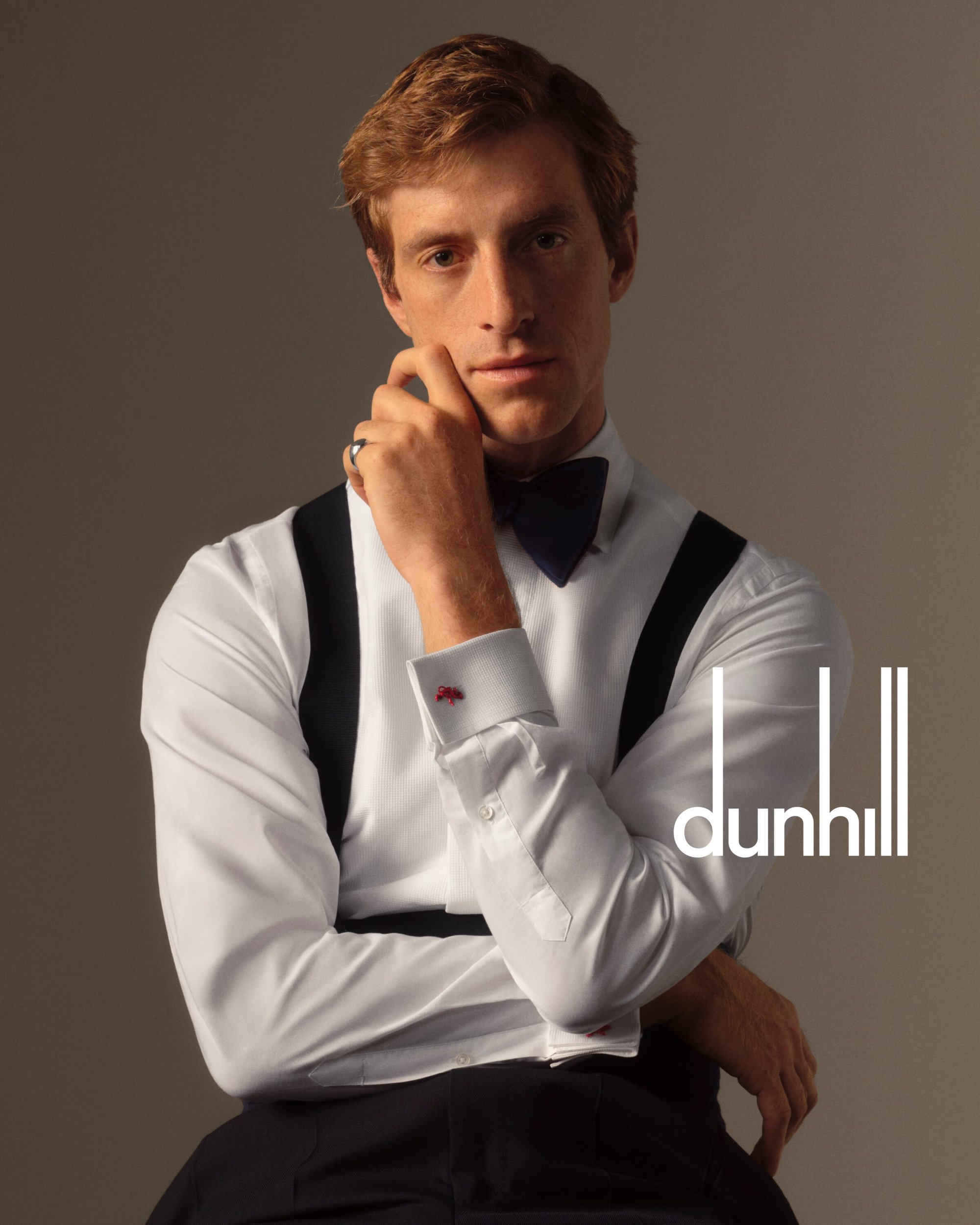 Portrait of a blond male model in formal wear and suspenders, gazing at the camera, with the Dunhill logo visible.