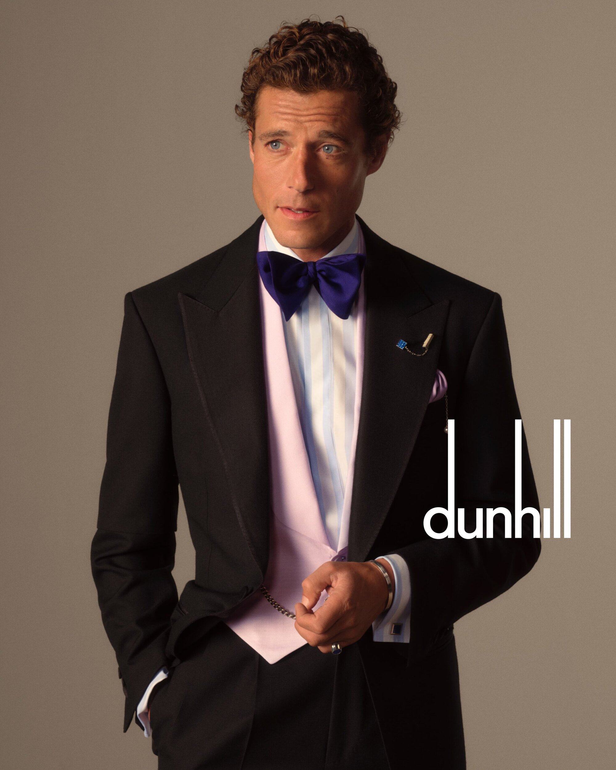 Male fashion model poses indoors wearing a tuxedo, vest, and bow tie, showcasing formal wear and luxury fashion in a studio setting.
