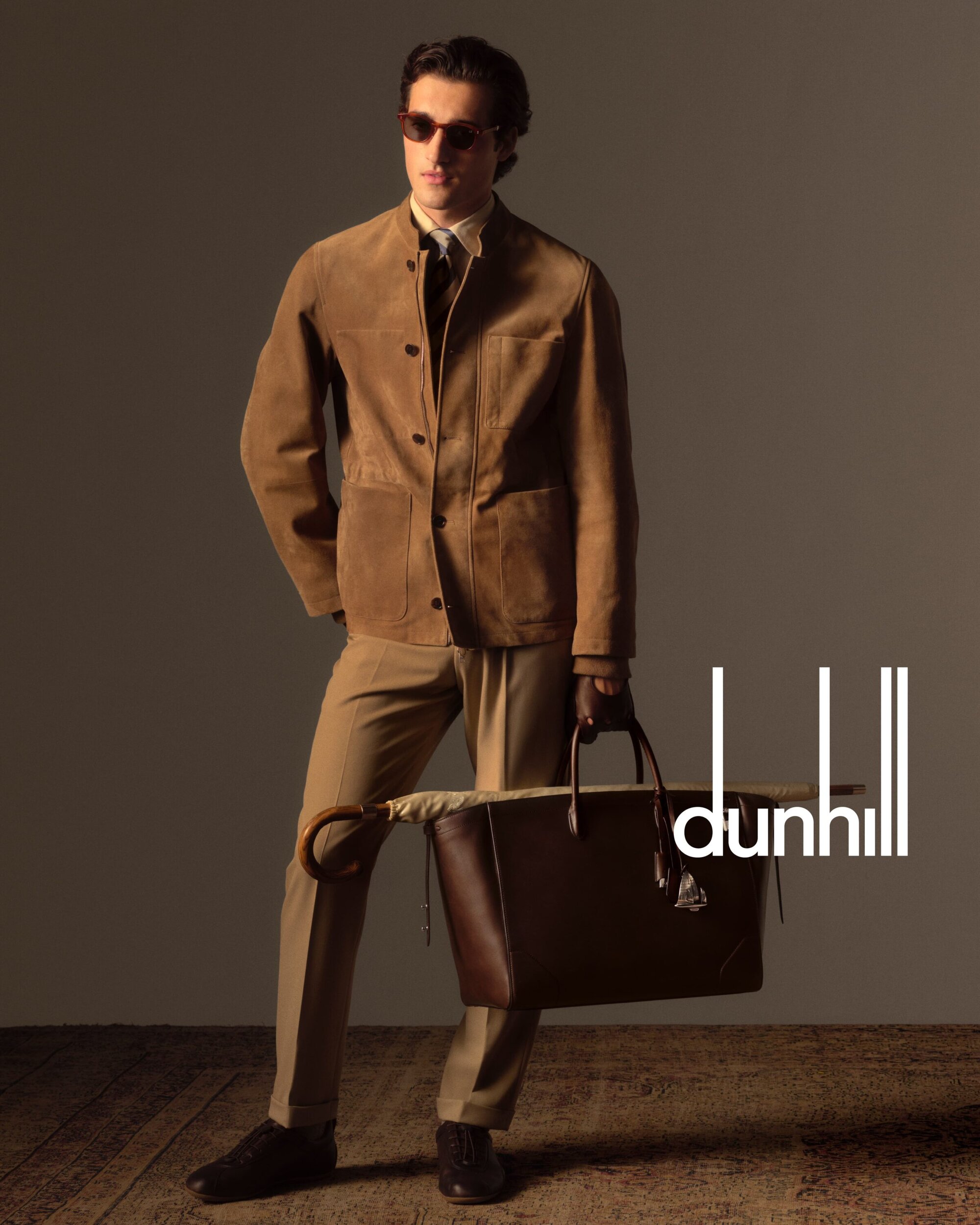 A man in a tan suit poses indoors with a brown leather bag, wearing sunglasses and a brown jacket in a fashion style