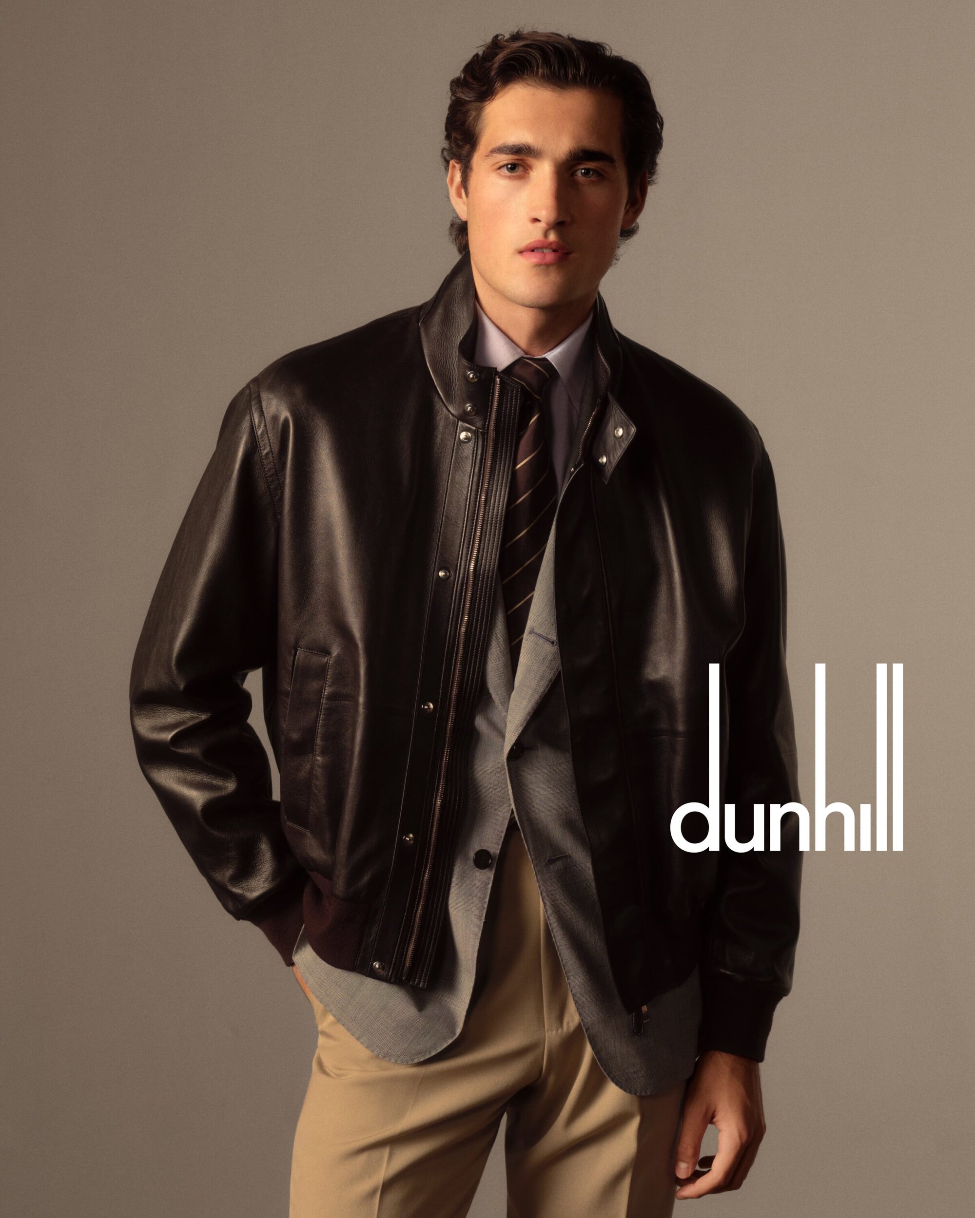 A fashionable adult male poses indoors in a brown leather bomber jacket, grey suit, patterned tie, and khaki pants with a neutral backdrop.