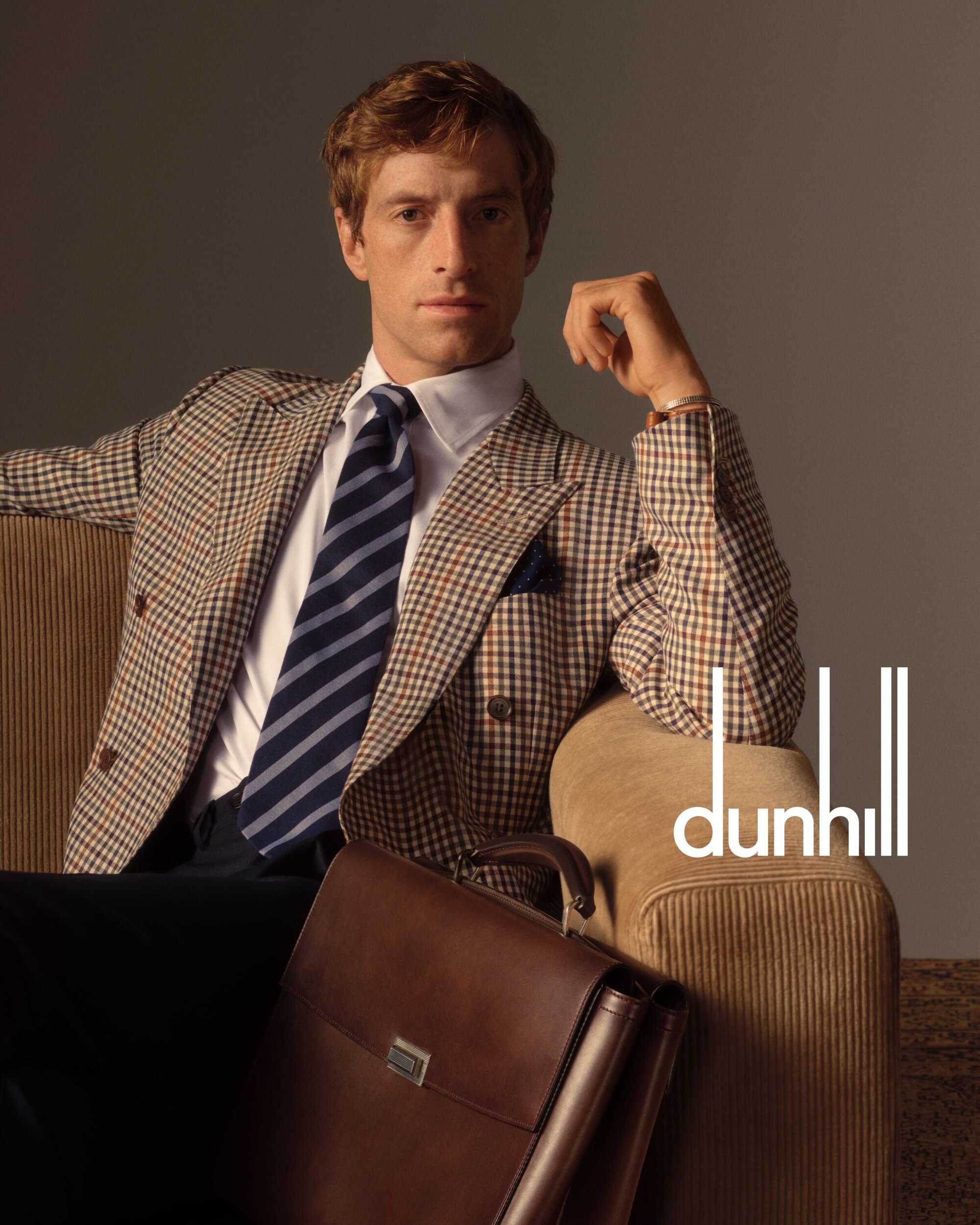 Portrait of a red-haired man in a blazer, white shirt, and striped tie, seated next to a brown leather briefcase and the Dunhill logo.