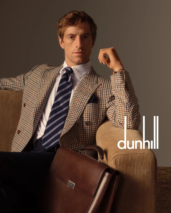 Man wearing a plaid blazer, white shirt, and striped tie seated next to a brown leather briefcase against a neutral background