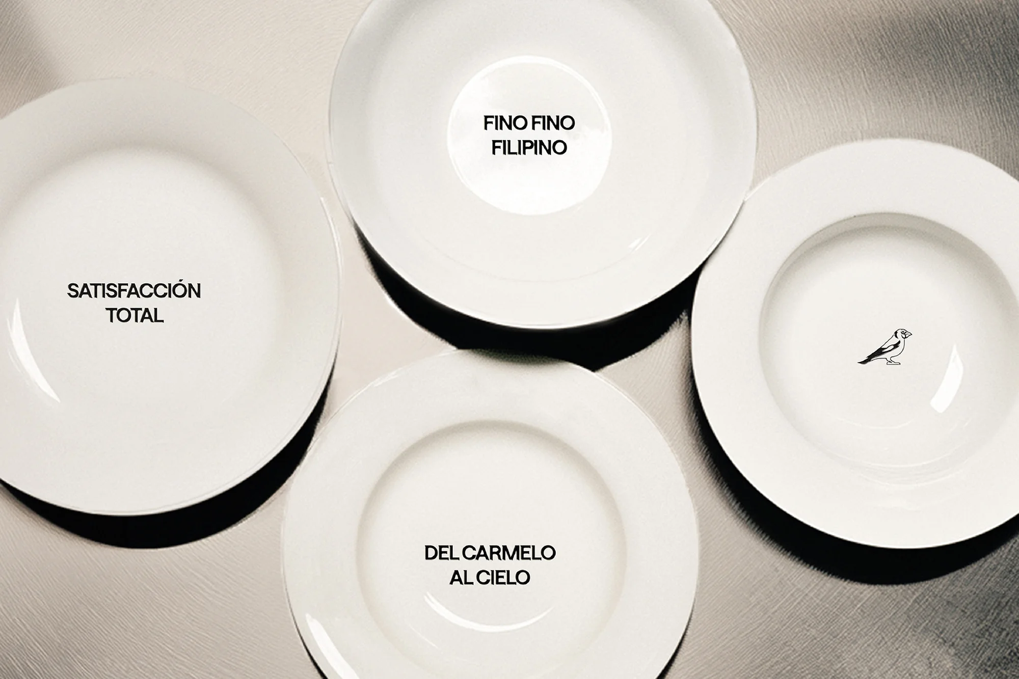 Overhead studio shot of four white, round plates on a neutral surface with black text inscriptions and a small line-art bird on one plate.