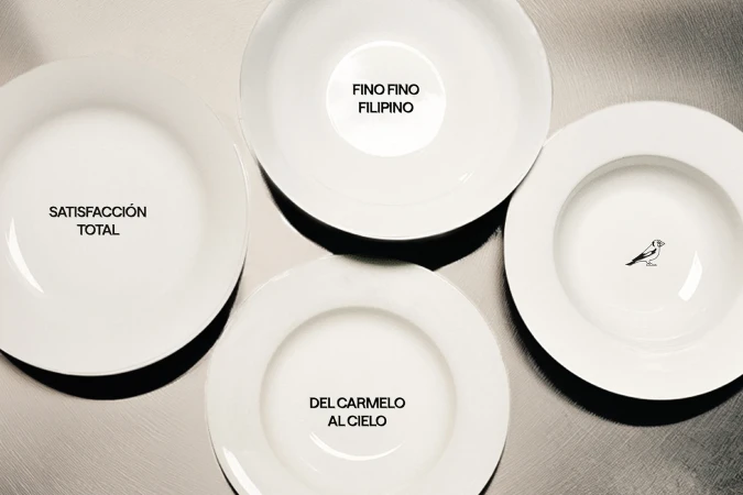 Overhead studio shot of four white, round plates on a neutral surface with black text inscriptions and a small line-art bird on one plate.