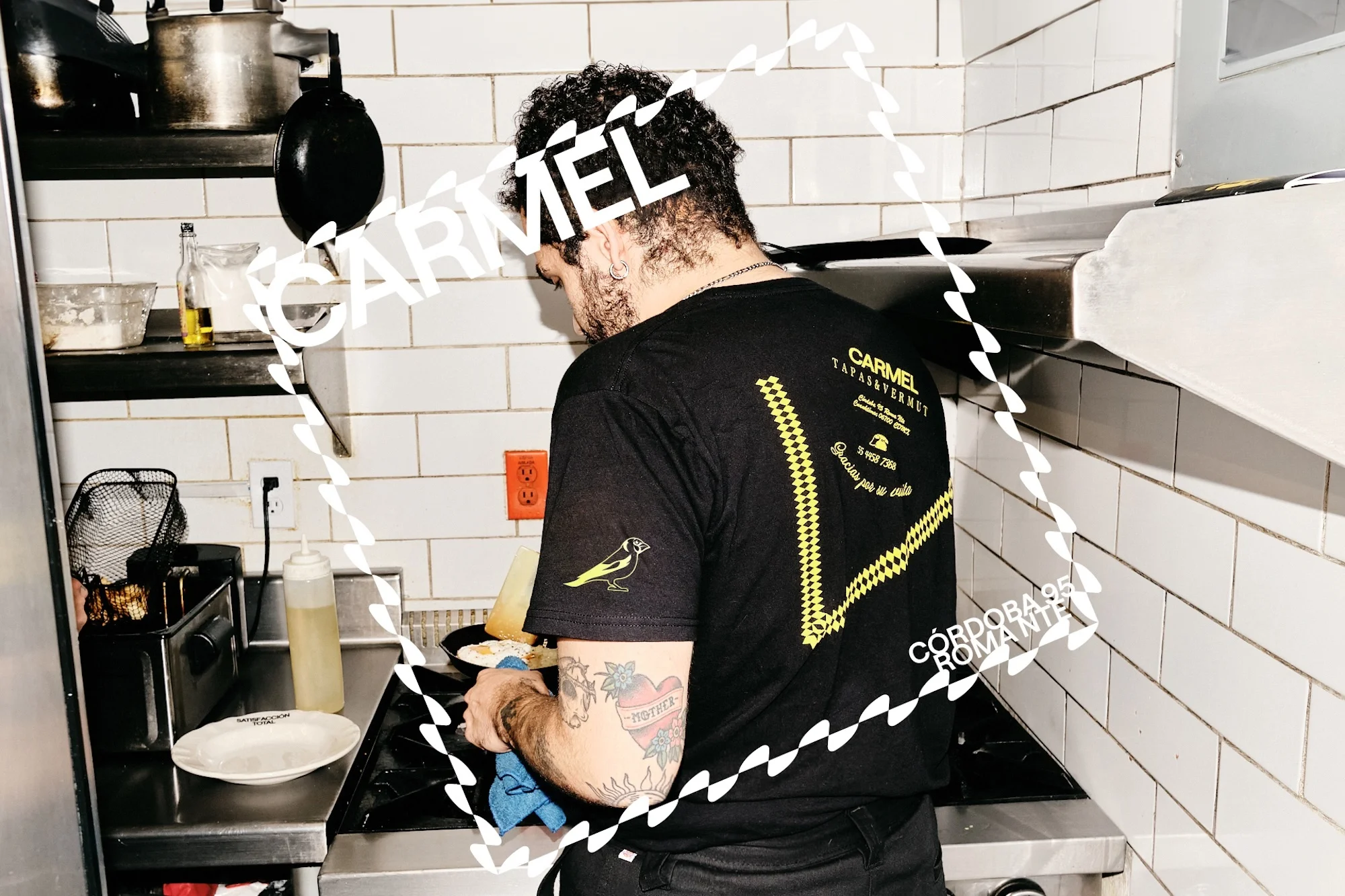 An adult male is cooking in a restaurant kitchen, wearing a black t-shirt with a logo, near a stove and metal cooking equipment.