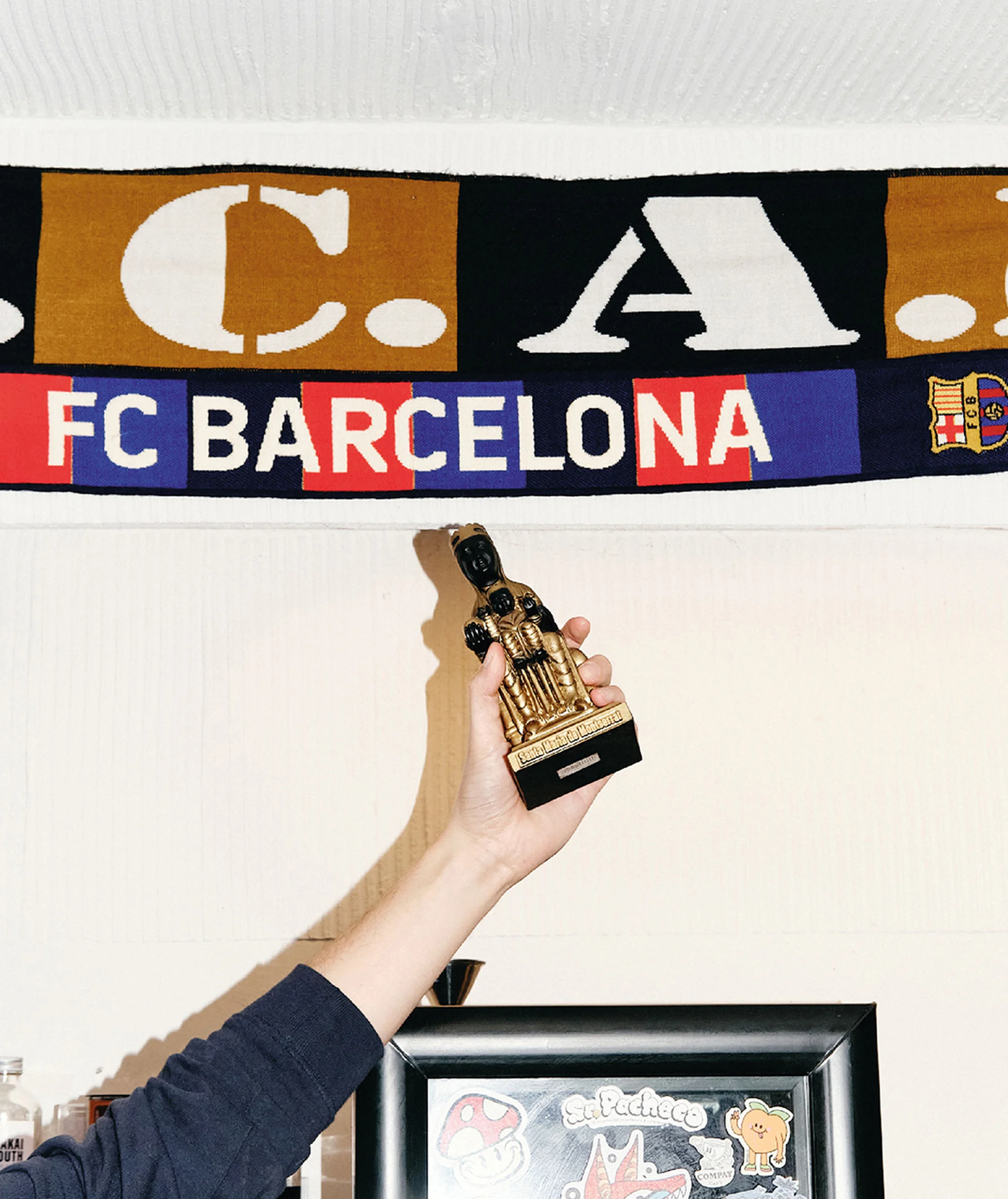 Close-up of a hand holding a golden statue, framed by an FC Barcelona scarf and framed artwork with stickers on a white wall.