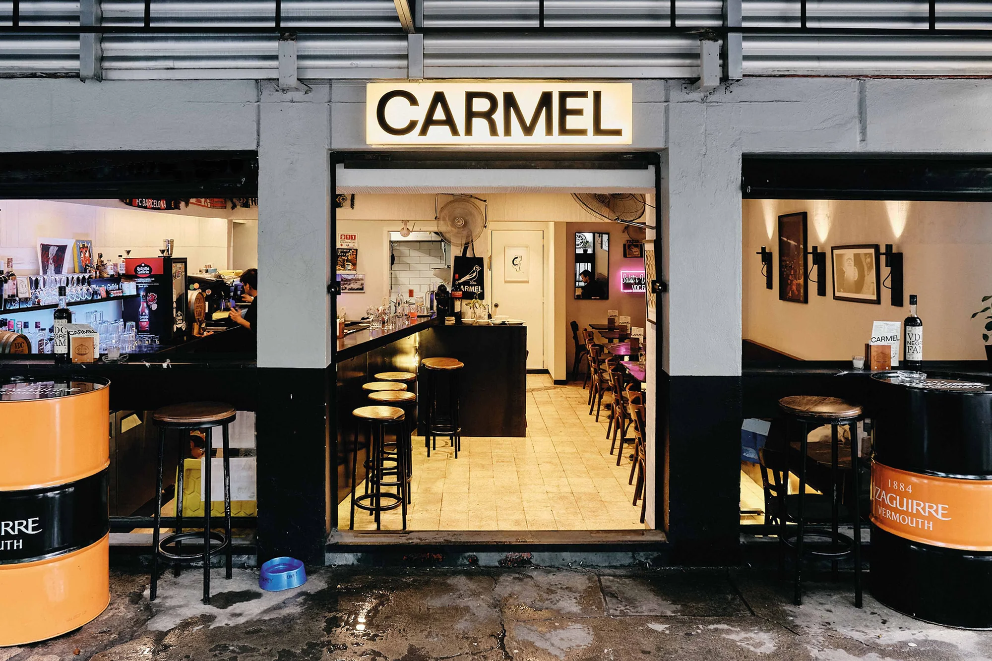 Warmly lit bar interior featuring stools at counter, liquor bottles, overhead sign reading "Carmel", and a partially visible dining area.