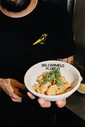 A person in a black t-shirt holding a white bowl of gnocchi garnished with fresh herbs for Carmel brand identity.