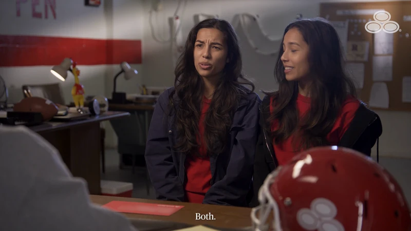 Two women converse in an office setting near a table with a football helmet and sports memorabilia, conveying a casual, indoor scene.
