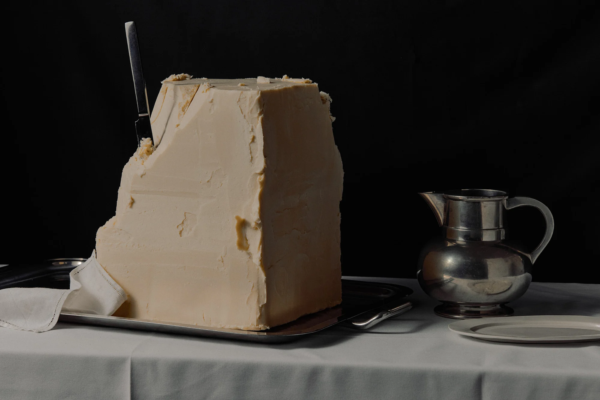 Still life shot of a large cut block of butter on a silver tray with a knife inserted, silver pitcher, and plate on a white tablecloth.