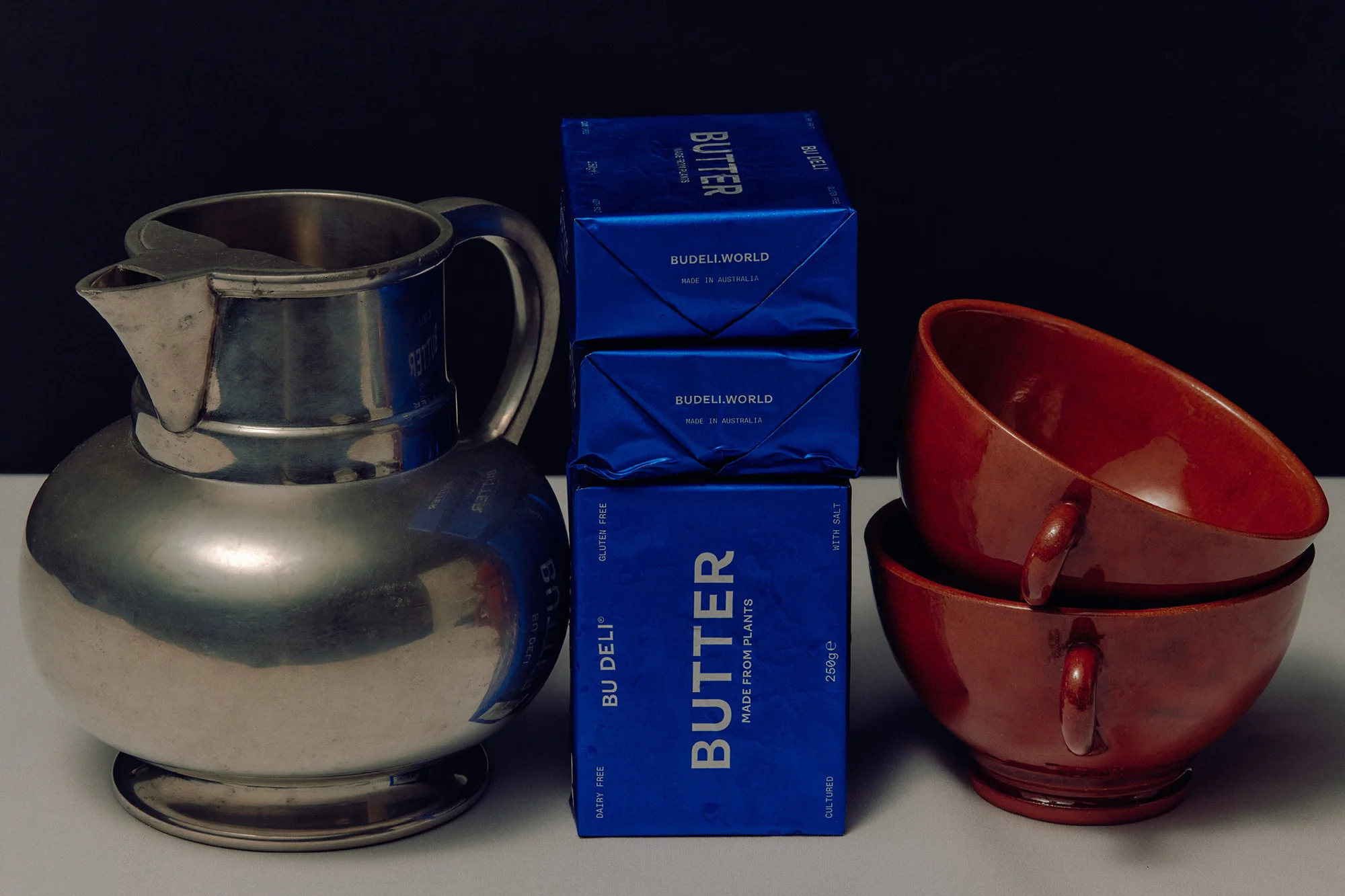 Still life featuring a silver pitcher, two stacked boxes of butter, and two red ceramic bowls against a dark background.