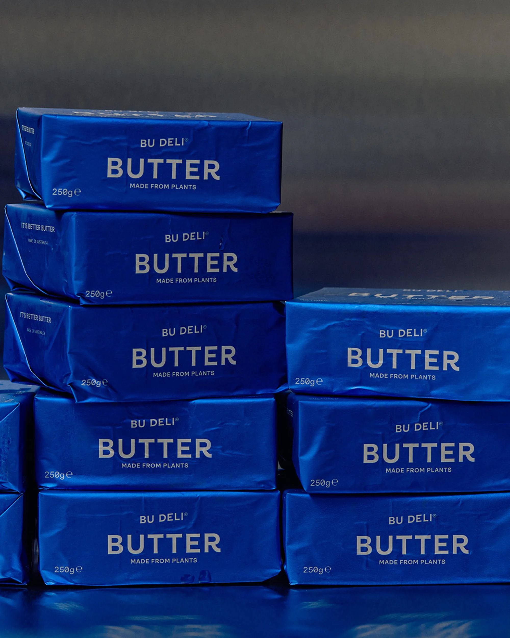 Stack of 'Bu Deli' brand vegan butter wrapped in blue packaging, displayed on a blue reflective surface against a dark background.