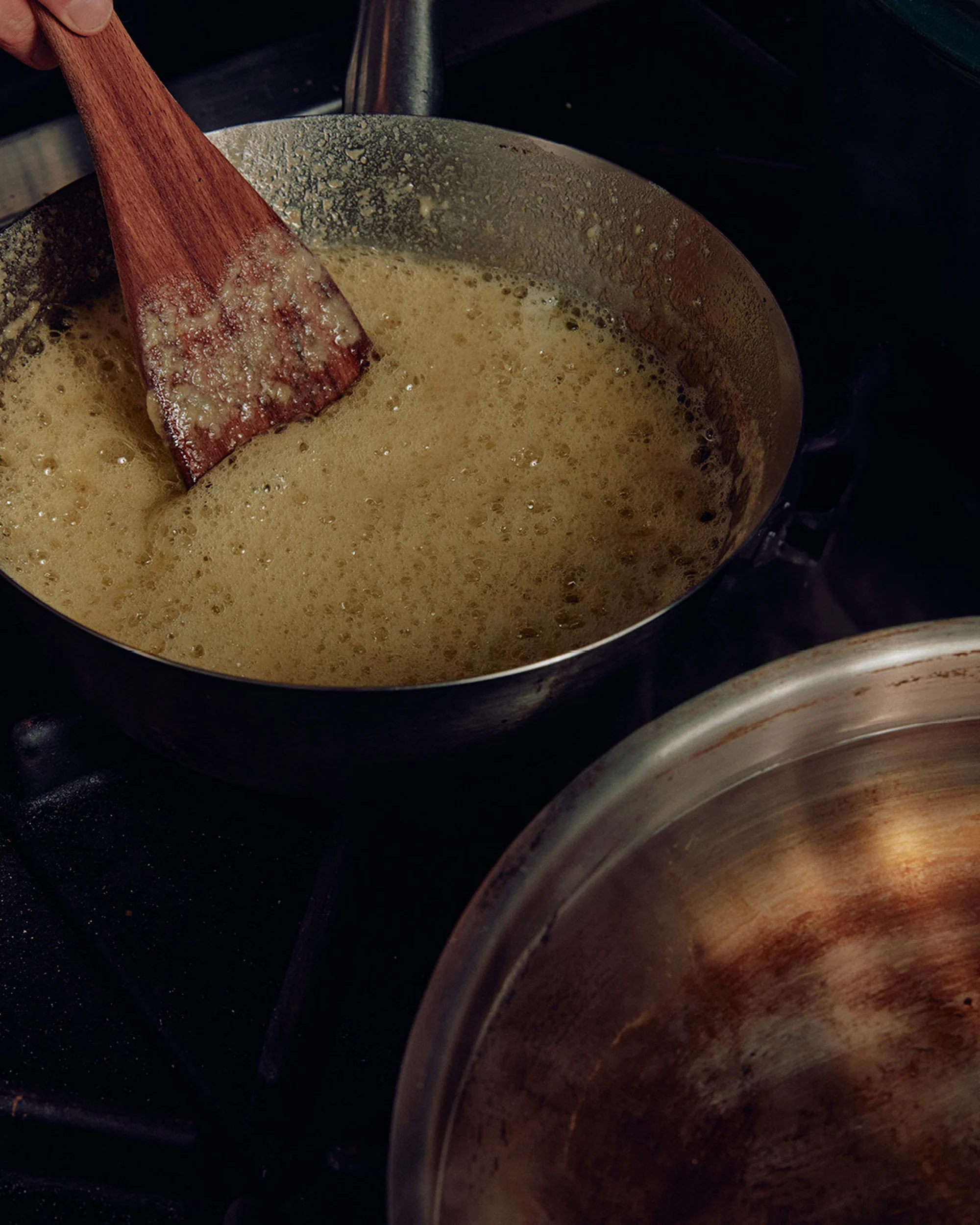Wooden spatula stirring a light yellow bubbling liquid in a metal pan on a black stovetop, another metal pan visible.