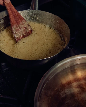 Wooden spatula stirring a light yellow bubbling liquid in a metal pan on a black stovetop, another metal pan visible.