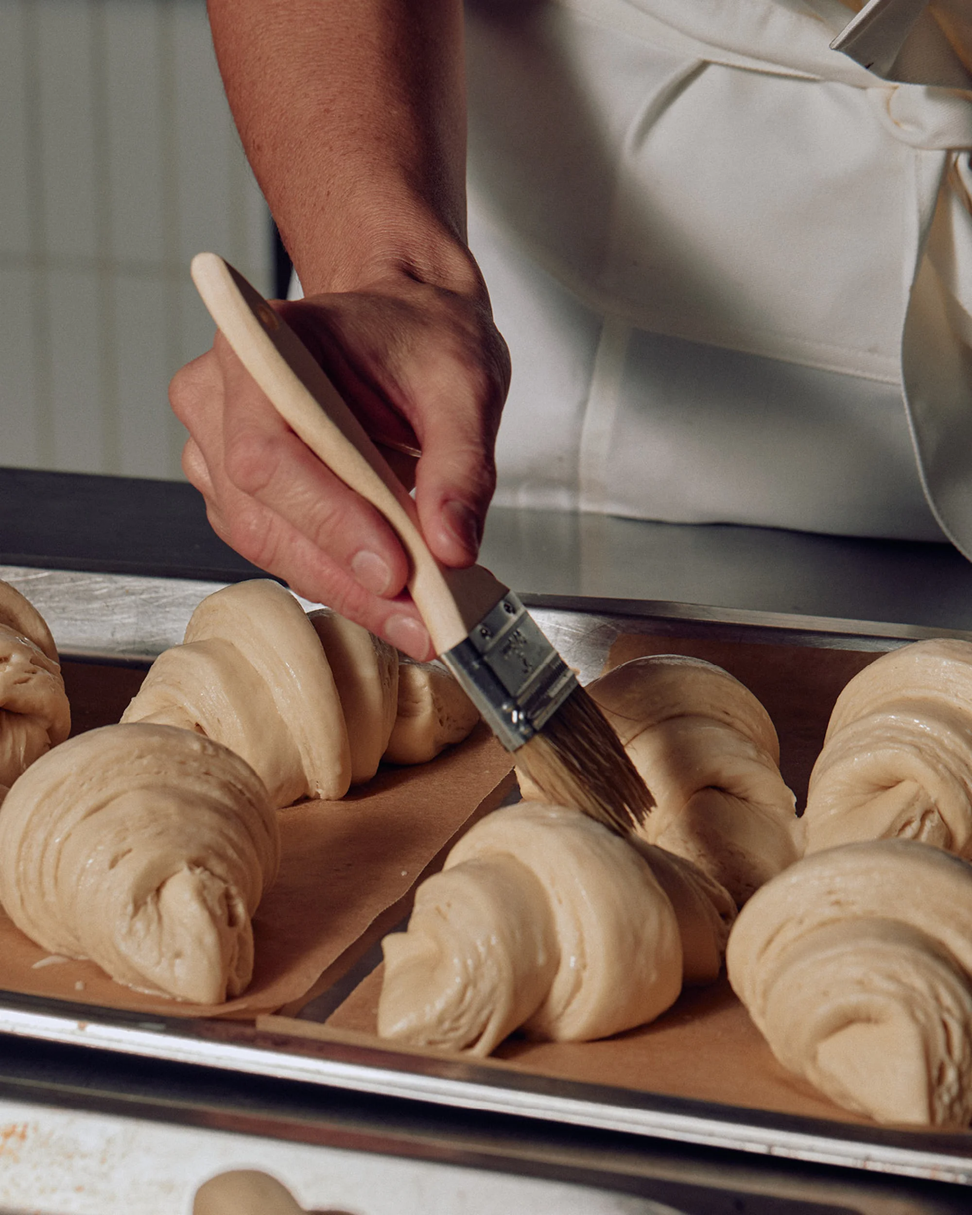 Close-up of a baker brushing raw croissants with an egg wash on a baking sheet in preparation for baking in a professional kitchen.