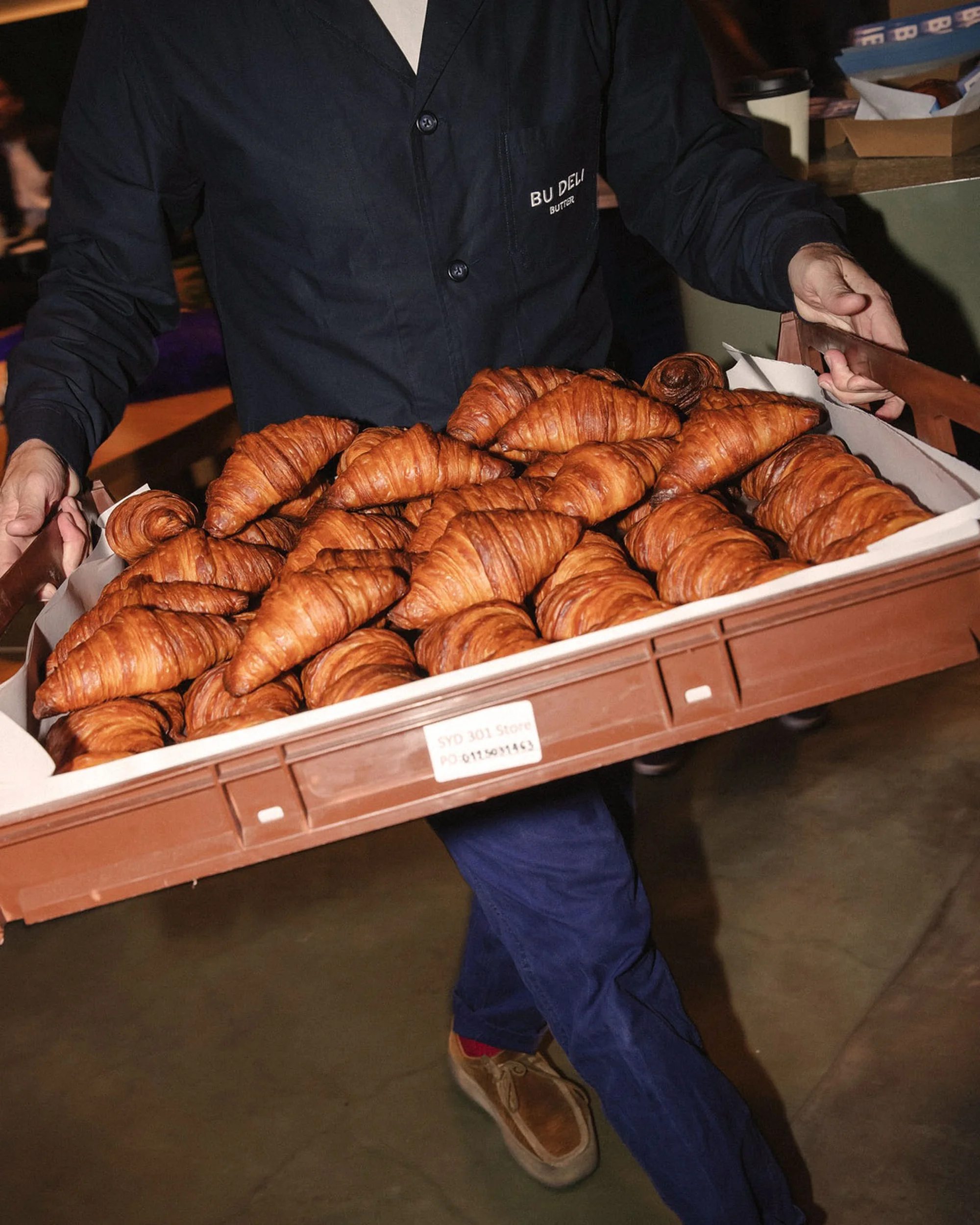 Person carries a tray overflowing with golden-brown croissants indoors, focusing on a close-up of the pastries and the carrier's hands.