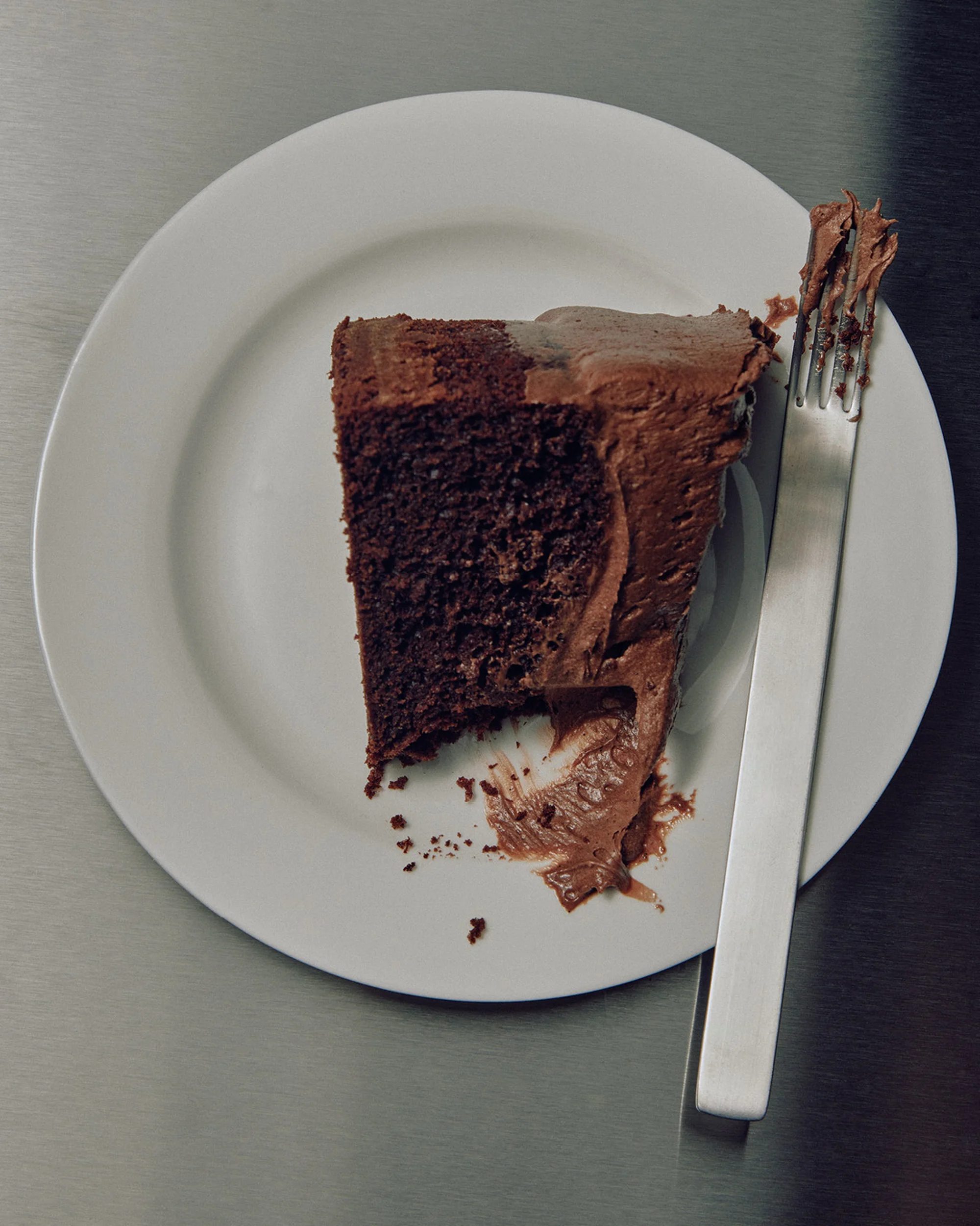 Overhead shot of a slice of chocolate cake with frosting on a white plate, accompanied by a silver fork, against a gray surface.
