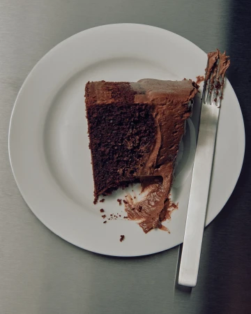 Overhead shot of a slice of chocolate cake with frosting on a white plate, accompanied by a silver fork, against a gray surface.