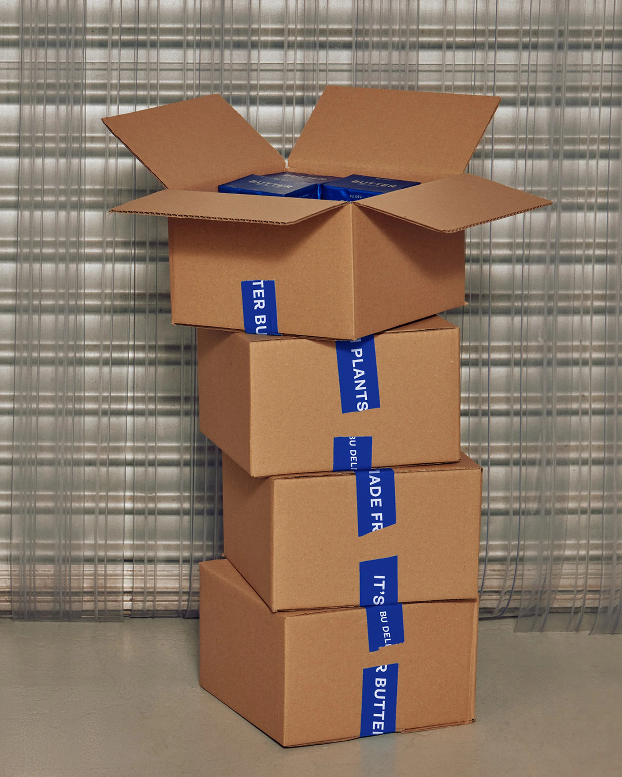 Stacked cardboard boxes, sealed with branded tape, suggest shipping or storage, with a closed metal shutter in the background.