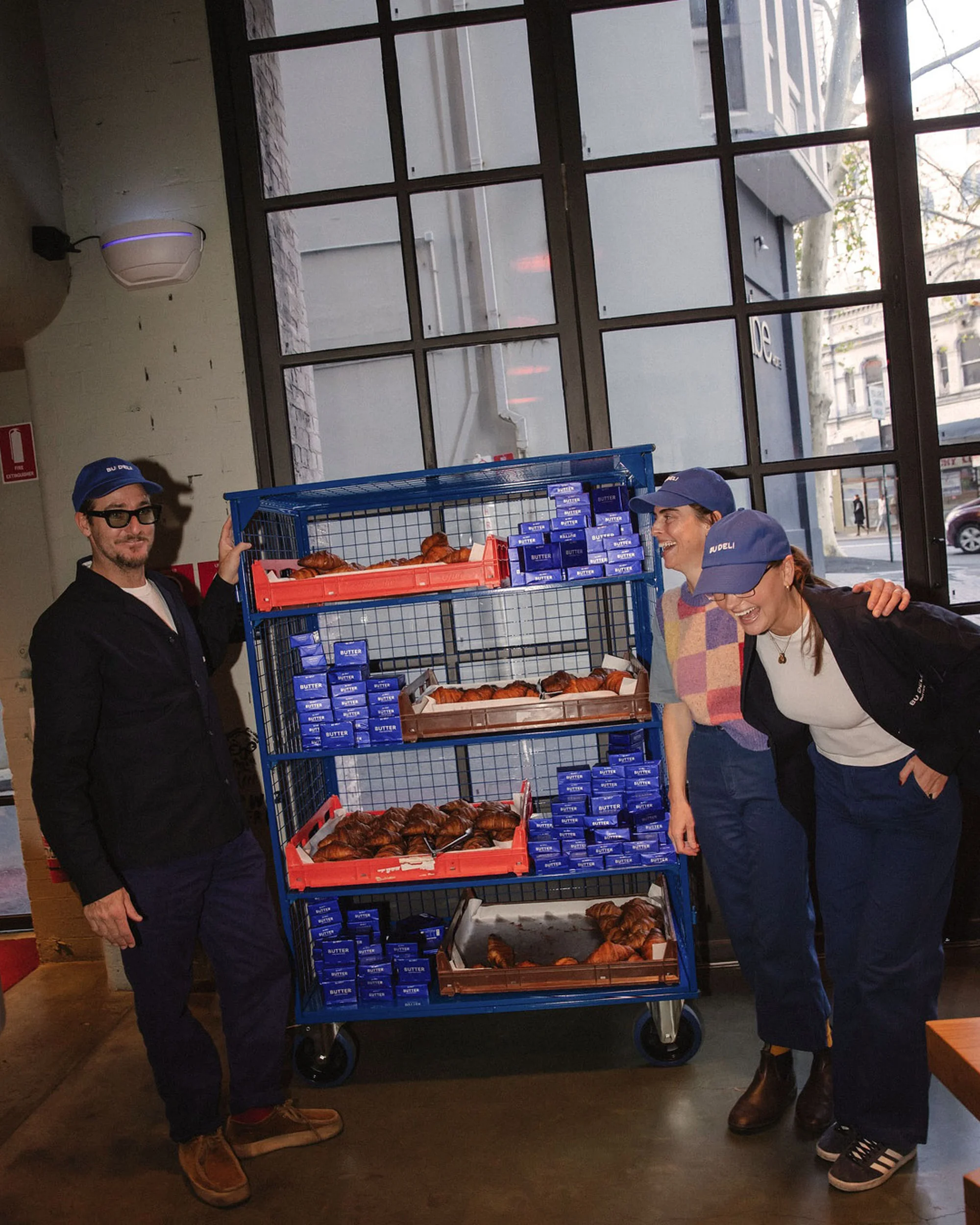 Group of three adults in matching hats standing with a metal cart filled with croissants and blue boxes in an indoor urban cafe setting.